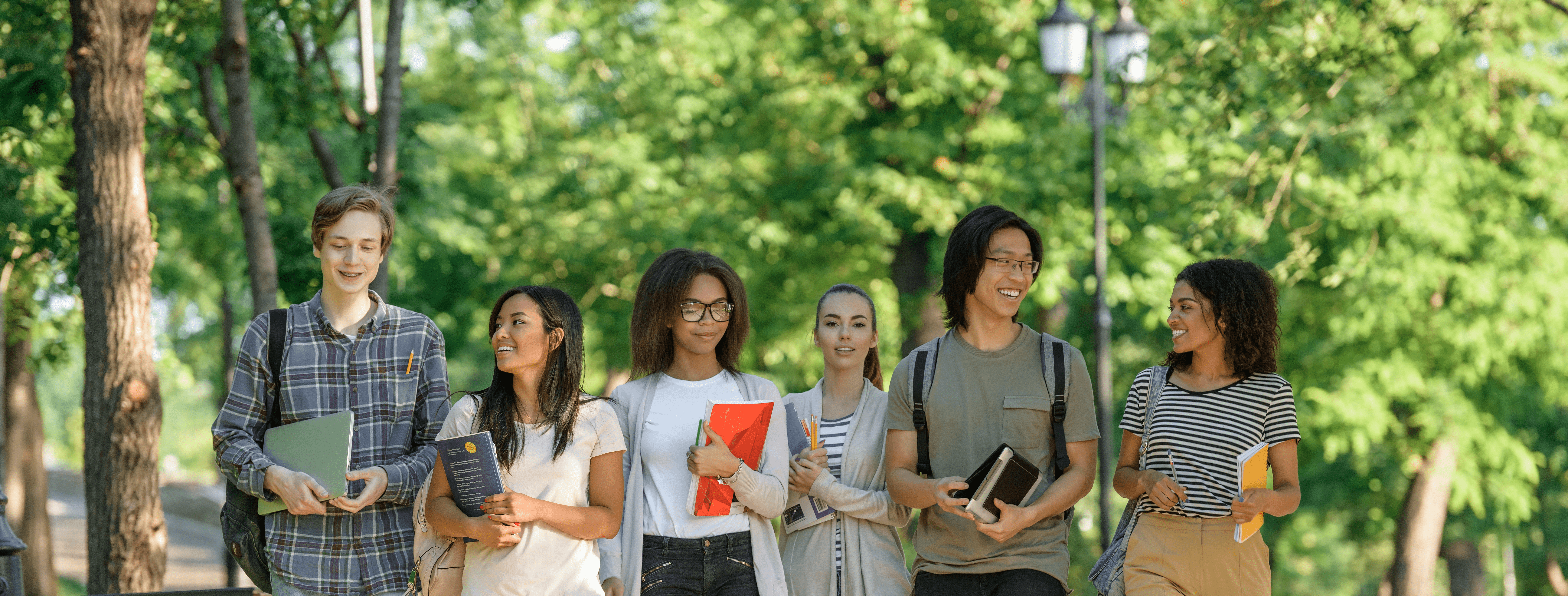 Un grup divers de studenți zambind și discutănd se plimbă, în linie, în aer liber, pe o alee însorită a campusului, mărginită de copaci înalți, luxurianți, într-o atmosferă luminoasă și plină de viață