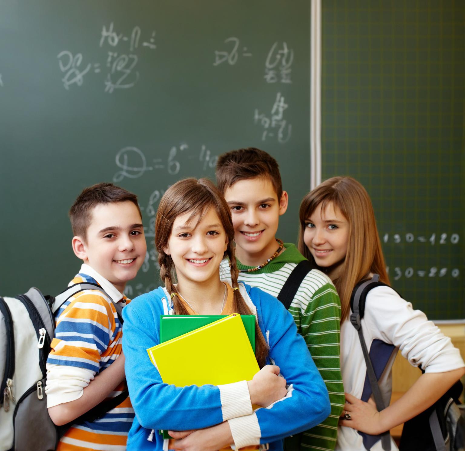 Un grup de elevi zâmbitori, cărând ghiozdane și caiete colorate, pozează în fața unei table scrise dintr-o sală de clasă într-o atmosferă veselă, academică