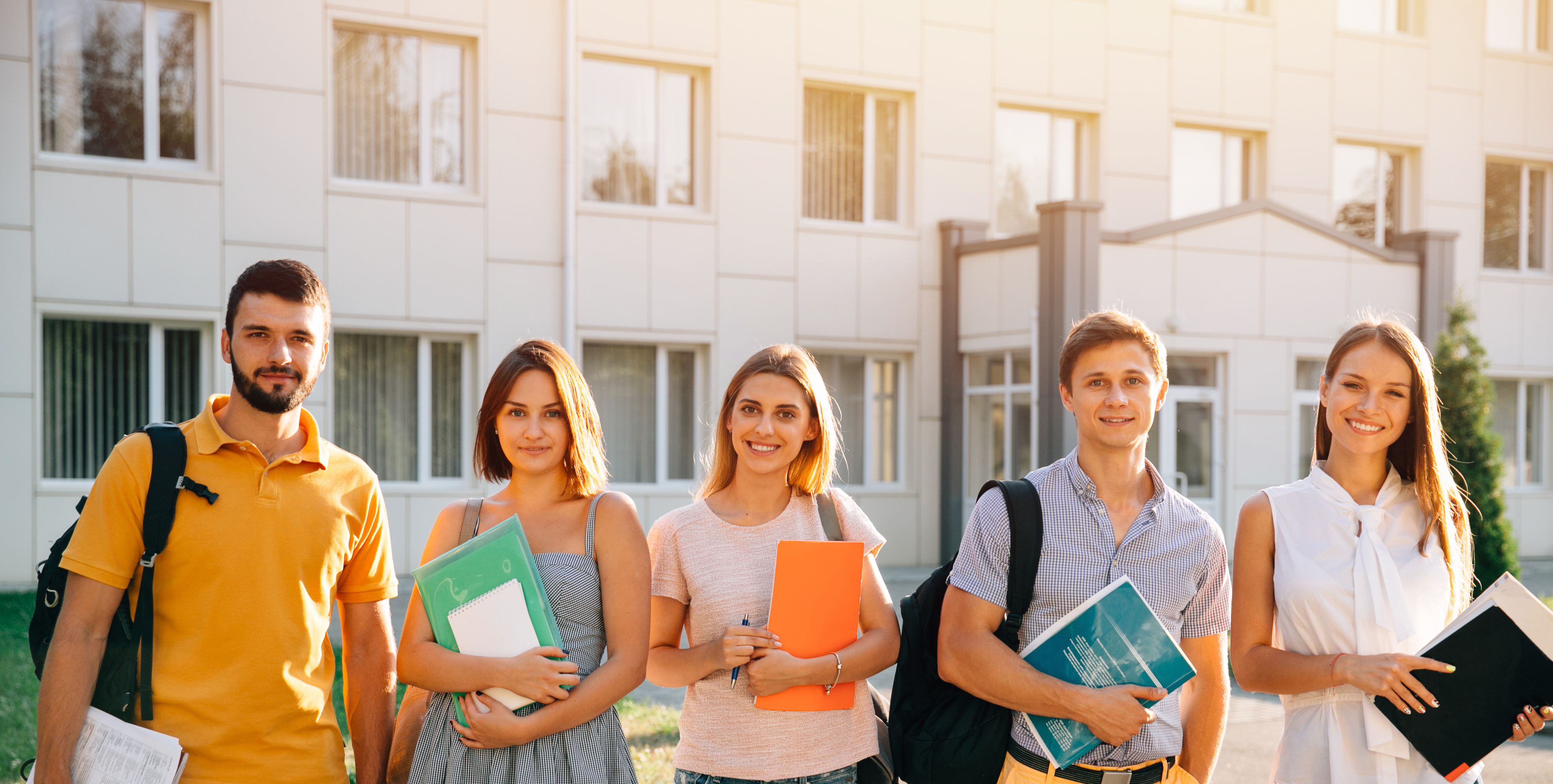 Un grup divers de studenți zambind, purtând rucsacuri ușoare și ținând în mâini cărți și caiete colorate, pozează, în linie, în fața unei clădiri universitare moderne, într-o atmosferă veselă și de studiu