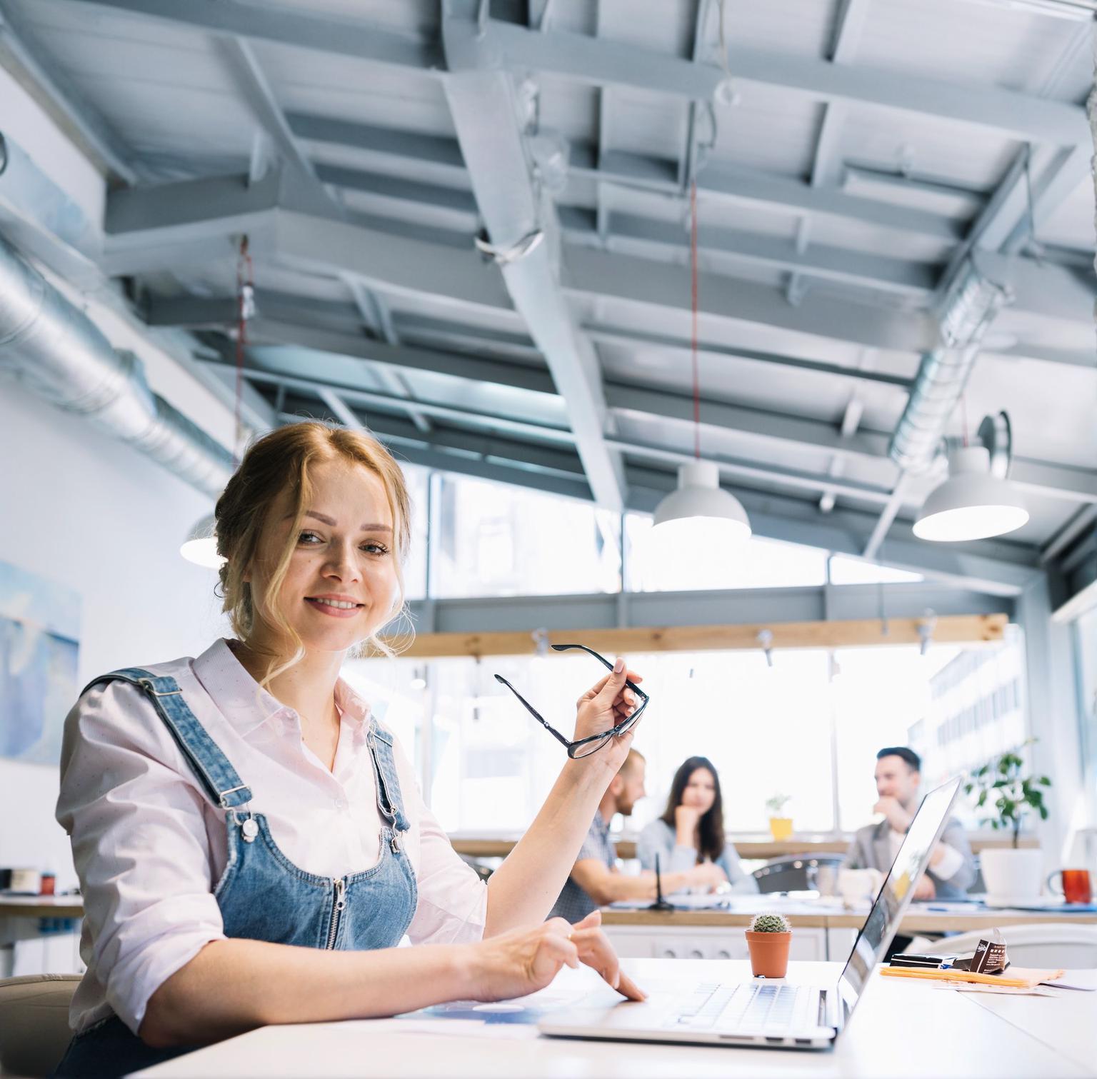 O studentă zâmbitoare, ținând în mână ochelarii, lucrează la un laptop într-un laborator modern, cu conducte de aer expuse în tavan, iar în fundal, estompați, sunt alți studenți care discută într-o atmosferă plină de viață