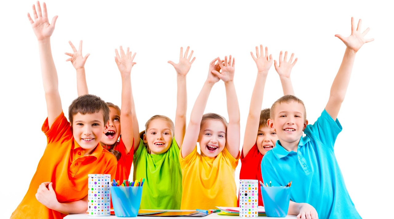 Un grup de elevi zâmbitori, în cămăși viu colorate, ridică mâinile cu entuziasm în spatele unei mese cu caiete colorate și suporturi cu creioane și culori, transmițând bucurie și entuziasm