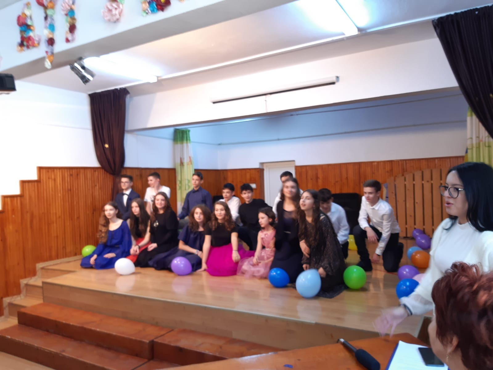 Grup de elevi asezați, cu baloane, pe scena Sălii de festivități a școlii