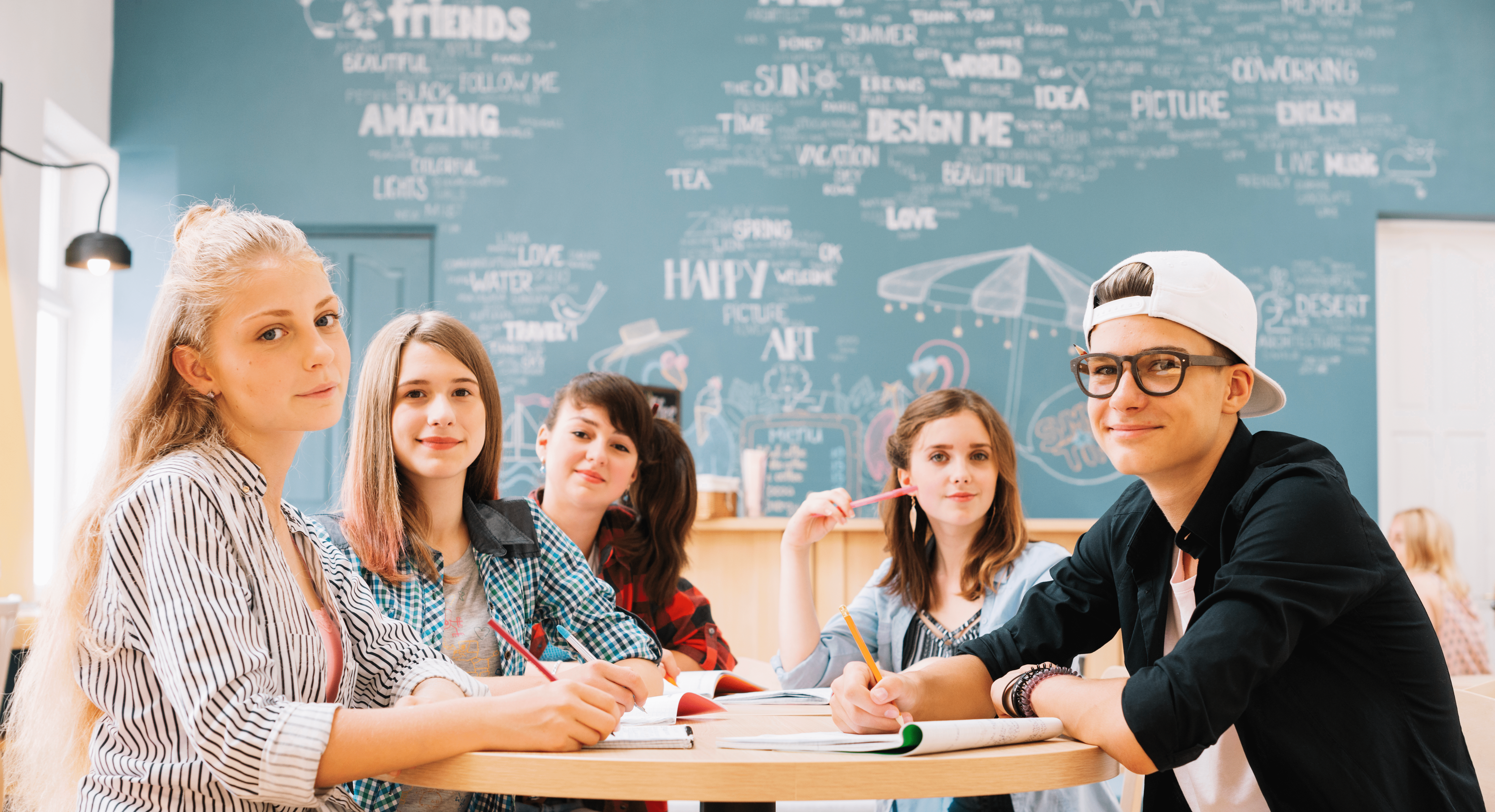 Un grup de elevi stau asezați în jurul unei mese rotunde, ținând în mână creioane colorate cu care scriu în caiete deschise, zâmbind și pozând, într-o cameră animată, iar în fundal este un perete albastru deschis pe care sunt scrise, cu alb, cuvinte motivaționale