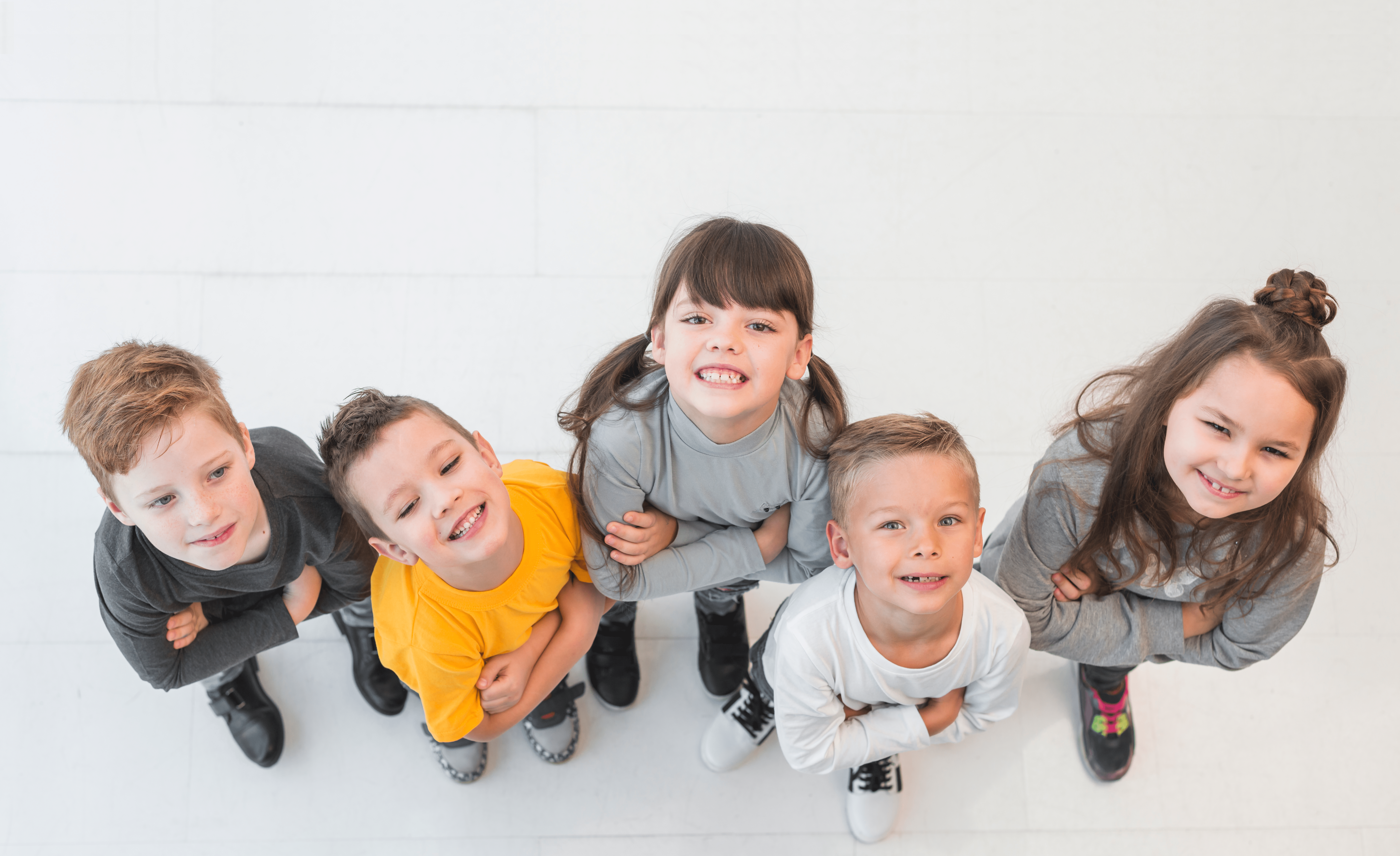 Un grup vesel de copii, purtând haine obișnuite în nuanțe de gri, galben și alb, stau aliniați pe o podea albă și privesc râzând direct la cameră dintr-o perspectivă de sus