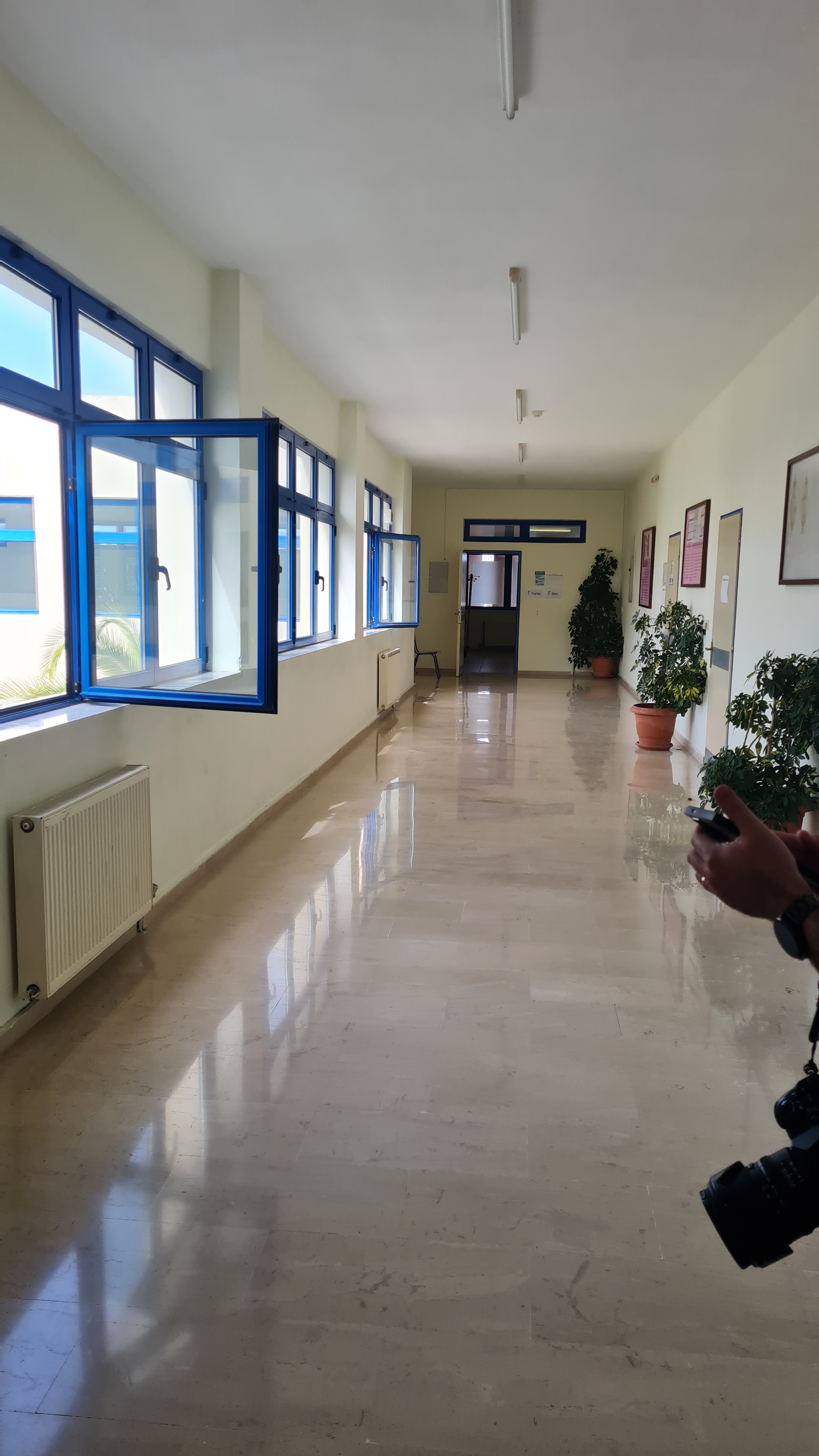 Un hol luminos al școlii, cu podea lucioasă din marmură, ferestre mari cu rame albastre deschise pe partea stângă, radiatoare albe sub ferestre și ghivece cu plante verzi aliniate pe partea dreaptă, cu pereți cu panouri informative, cu o ușă cu ramă albastră către o altă încăpere, în fundal, iar în colțul din dreapta jos apare parțial o cameră foto ținută de o persoană