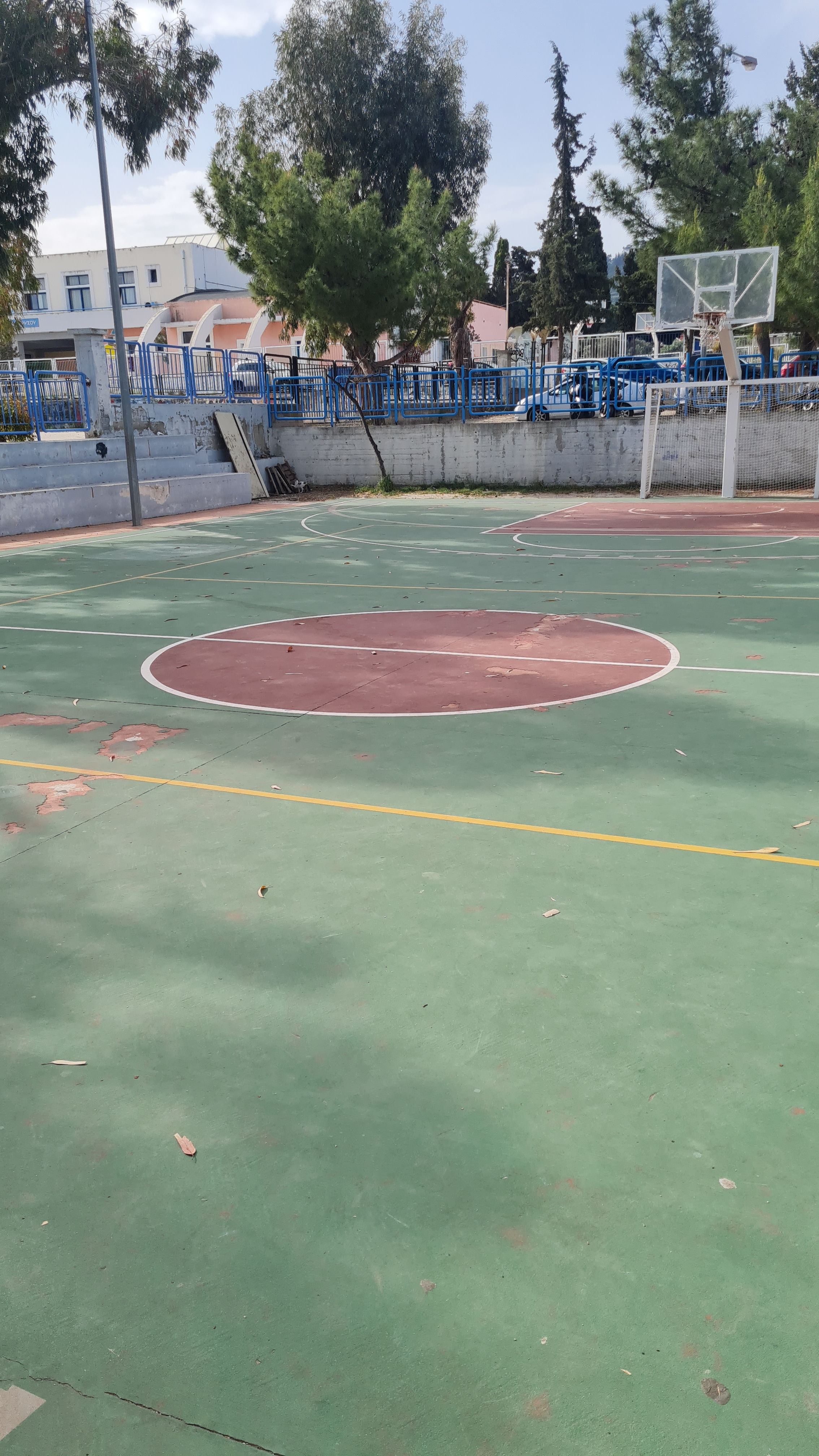 Un teren de baschet/handbal/fotbal/volei în aer liber cu suprafață verde și zone circulare maro, vizibil uzat și decojit, încadrat de gard albastru metalic, panou de baschet transparent și tribune de beton, iar în fundal sunt clădiri școlare, copaci înalți și cer parțial înnorat