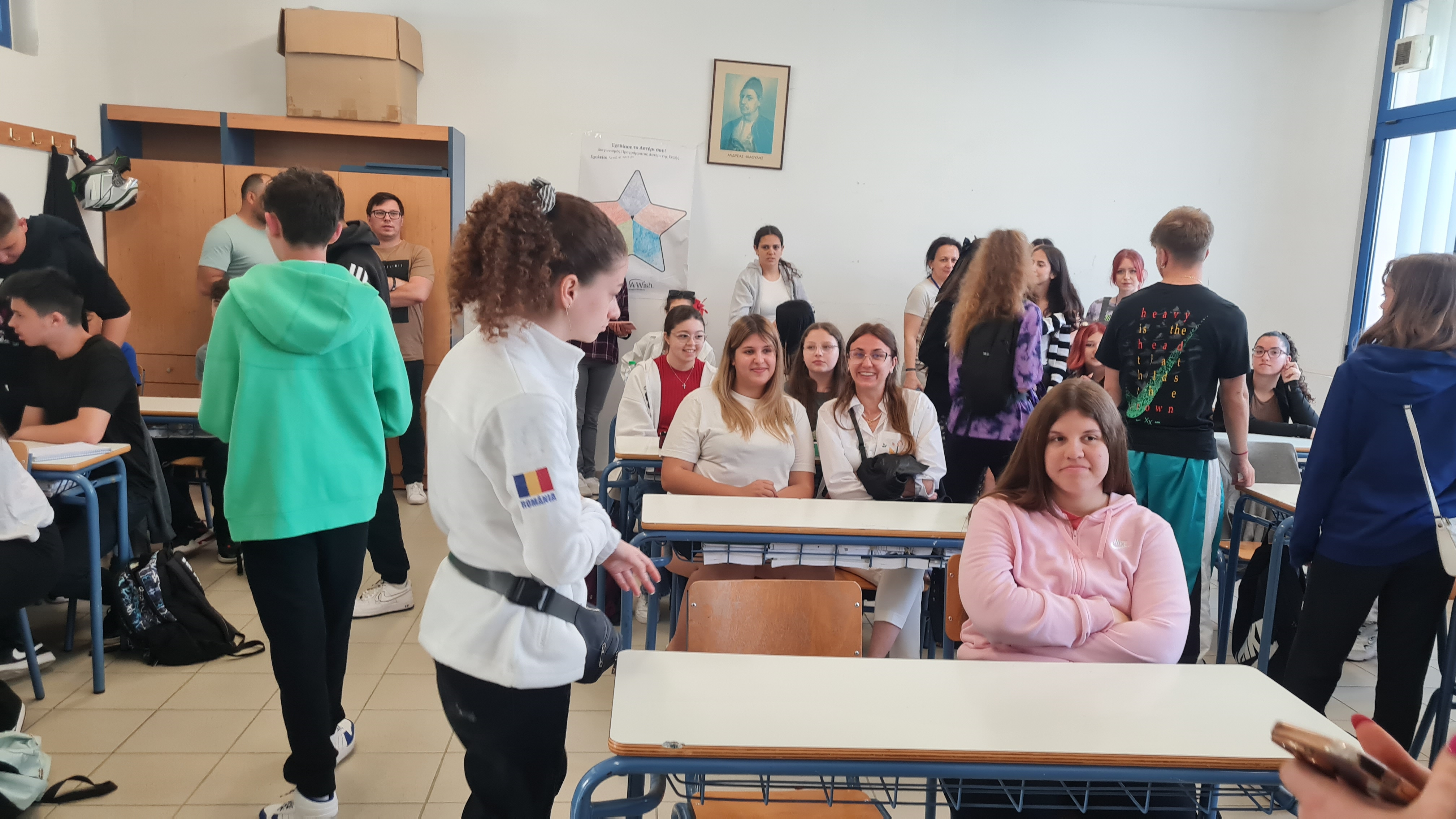 O sală de clasă aglomerată cu numeroși elevi adolescenți care stau la bănci sau discută în picioare, într-o atmosferă de pauză școlară animată, în prim-plan fiind o fată îmbrăcată în hanorac roz care stă așezată în bancă, cu brațele încrucișate, în centru fiind o elevă în bluză albă cu steagul României pe mânecă ce vorbește cu colegii, iar pe pereți sunt afișe educaționale, un portret și dulapuri din lemn albastru și maro deschis