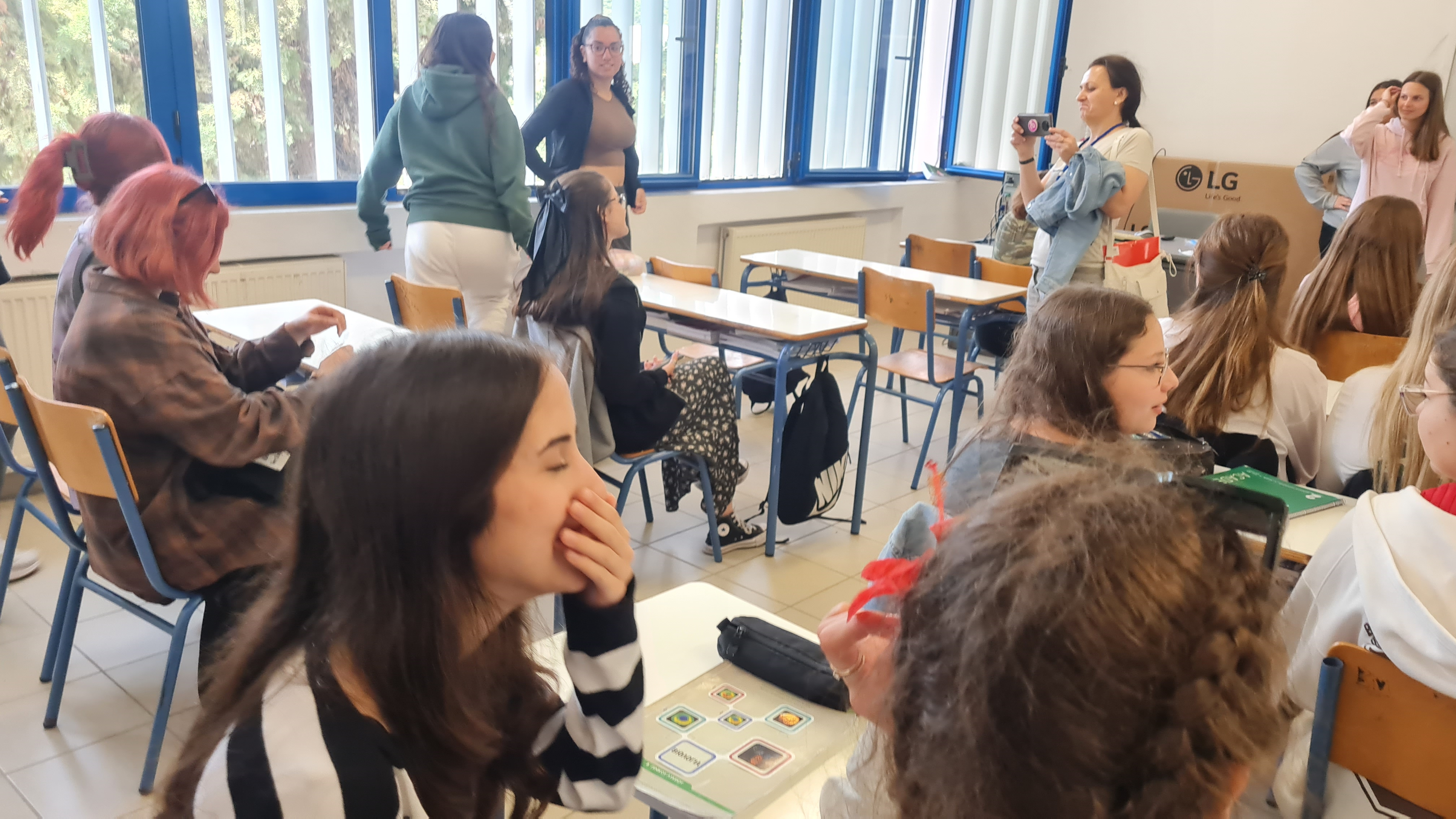 O sală de clasă luminoasă, cu ferestre mari, cu rama albastră, plină de elevi adolescenți așezați în bănci maro deschis, discutând și râzând, într-o atmosferă relaxată de activitate educațională, un cadru didactic fotografiază grupul, în prim-plan fiind câteva eleve cu păr roșcat și păr lung șaten care stau în bănci, pe care sunt autocolante colorate cu imagini tip joc educativ