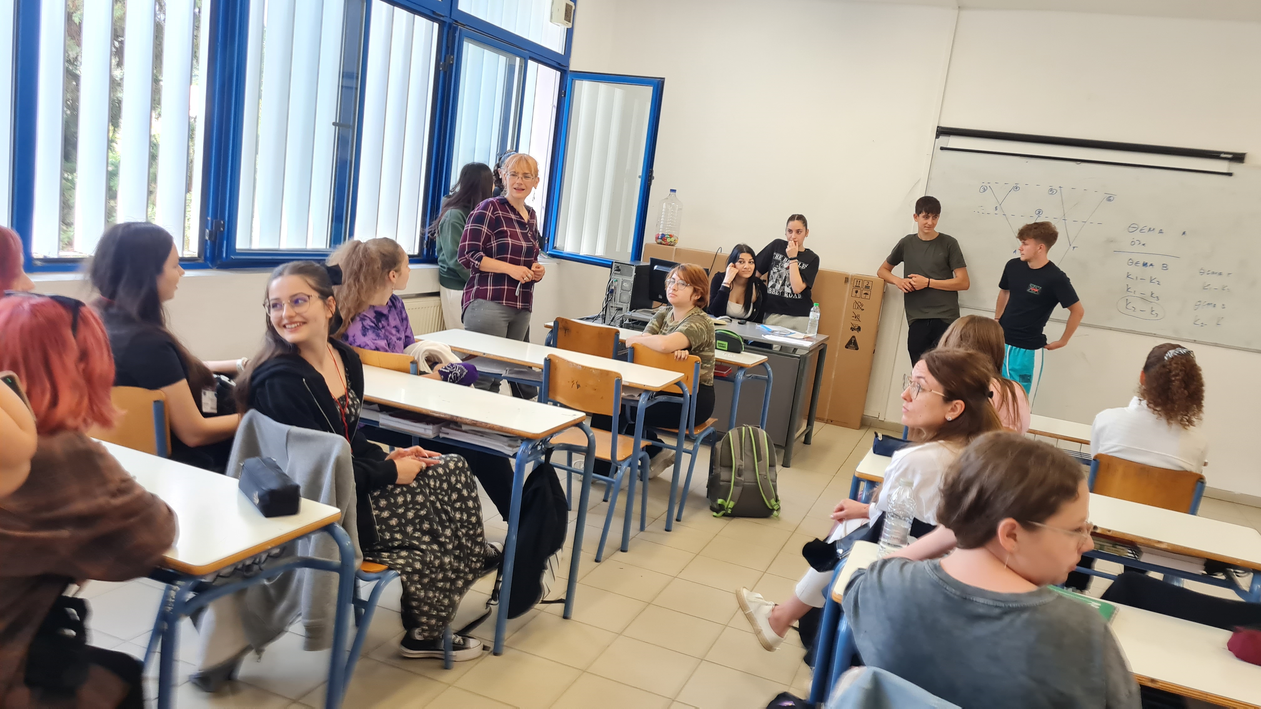 O sală de clasă luminoasă, cu ferestre mari, cu rama albastră, plină de elevi adolescenți aşezaţi la bănci maro deschis, discutând și zâmbind, într-o atmosferă de activitate educațională și prezentare în grup, un cadru didactic în cămașă în carouri stă în fața clasei, trei elevi stau lângă tabla albă pe care se văd diagrame și formule scrise, iar în partea dreaptă, în fața clasei, se află o catedră cu computer, pe care sunt caiete, pliante de prezentări și o sticlă cu apă