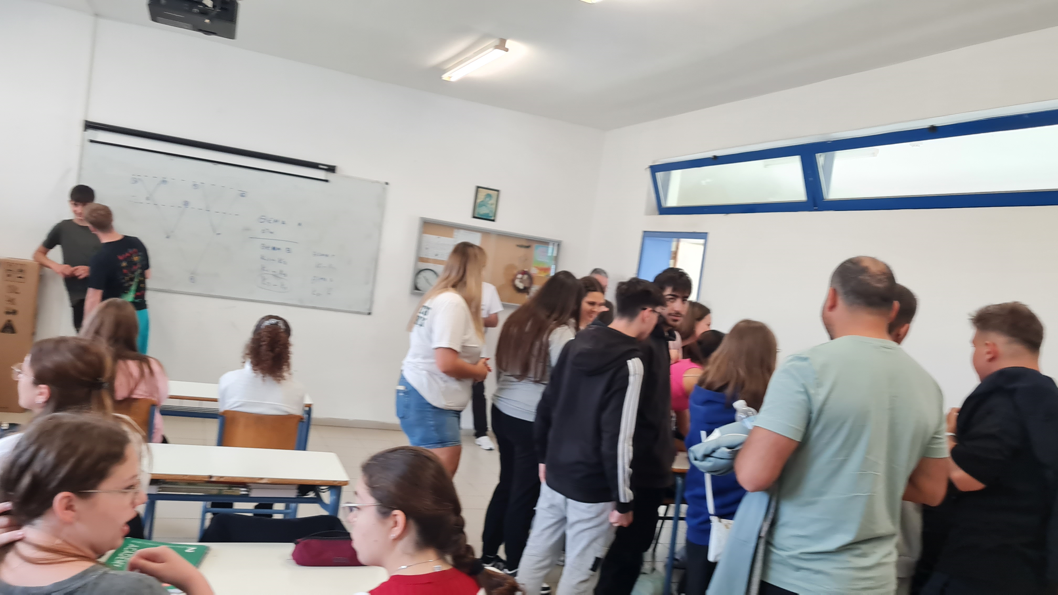 Elevi și adulți sunt adunați, într-o atmosferă de activitate participativă și mediu educațional, într-o sală de clasă luminoasă, cu ferestre cu rama albastră și mobilier școlar simplu, un grup stând în picioare în partea dreaptă și discutând, în timp ce alți elevi stau în bănci, iar în fundal este o tablă albă cu diagrame și calcule scrise