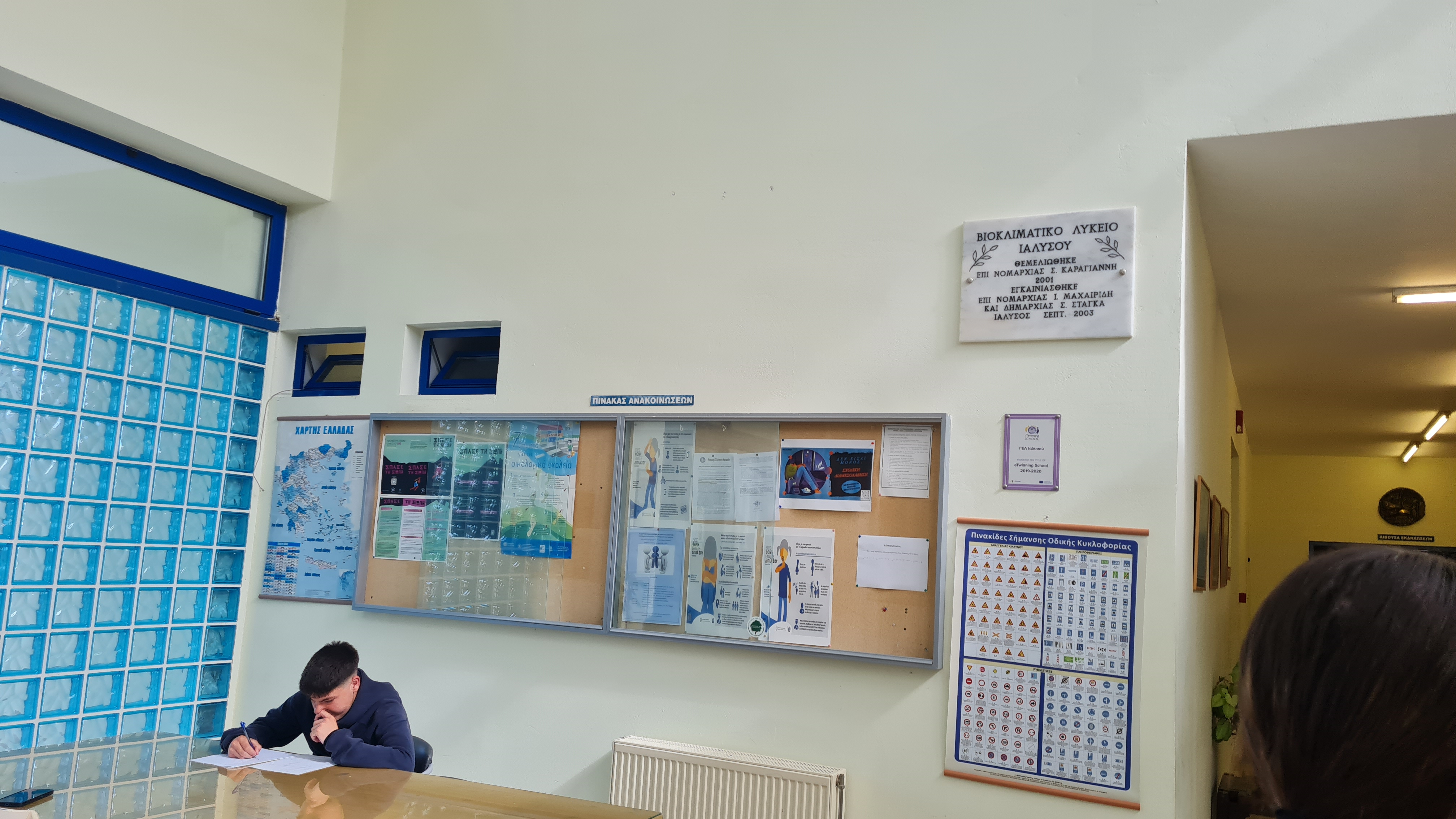 Într-un hol cu un perete din panouri groase de sticlă albastră transparentă si cu ceilalți pereți deschiși la culoare, pe care se află o placă de inscripție inaugurală ce indică, în limba greacă, „Liceul Bioclimatic din Ialyssosun”, un panou de anunțuri cu afișe educaționale, o hartă a Greciei, afișe cu instrucțiuni de postură corectă și informații administrative, precum și un poster cu semne rutiere, un elev stă la o masă lungă din lemn scriind pe niște foi albe de hârtie, iar în dreapta este un alt coridor cu iluminare fluorescentă albă
