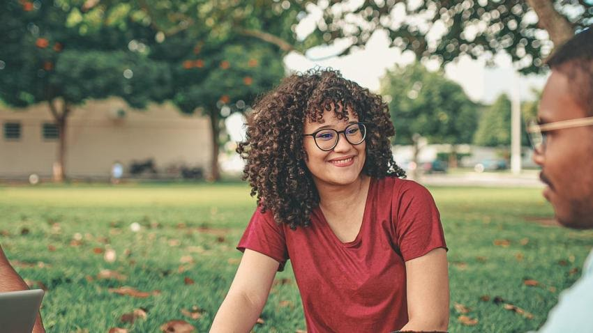 O persoană cu părul creț și ochelari zâmbește în timp ce stă pe iarba unui parc, purtând o cămașă roșie și vorbind cu o altă persoană, arătând o dispoziție prietenoasă