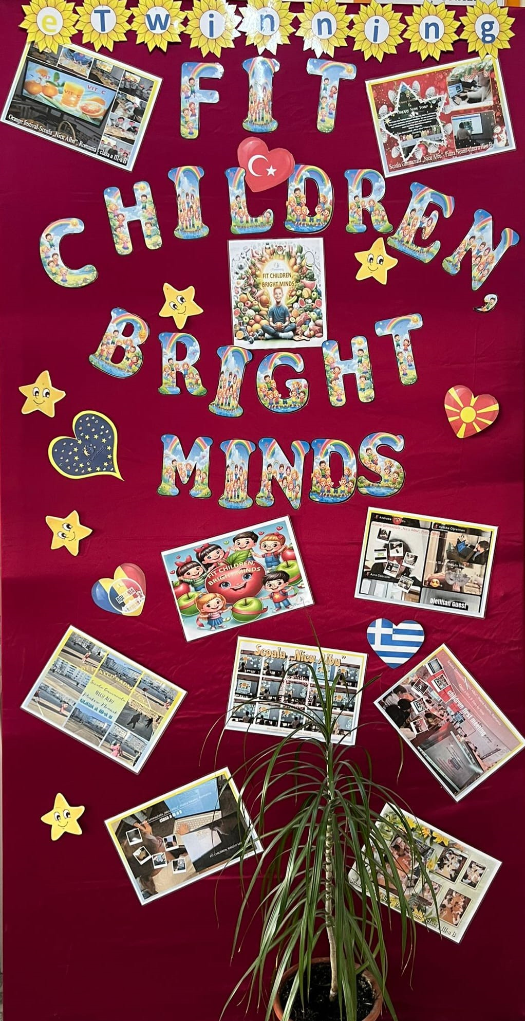 Panou decorativ cu fundal roșu, decorat cu flori tip floarea-soarelui, stele și inimi, inclusiv inimi cu steaguri ale diferitelor țări, pe care este afișat textul, în limba engleză, „eTwinning FIT CHILDREN, BRIGHT MINDS” (COPII ÎN FORMĂ, MINȚI STRĂLUCITOARE) realizat din litere colorate cu ilustrații de copii, în jurul textului sunt expuse mai multe fotografii și colaje care prezintă copii implicați în activități educaționale, ateliere și proiecte școlare, în centru se află un afiș cu tema alimentației sănătoase și a copiilor fericiți, iar jos, în fața panoului, este un ghiveci cu o plantă verde ce acoperă parțial câteva fotografii