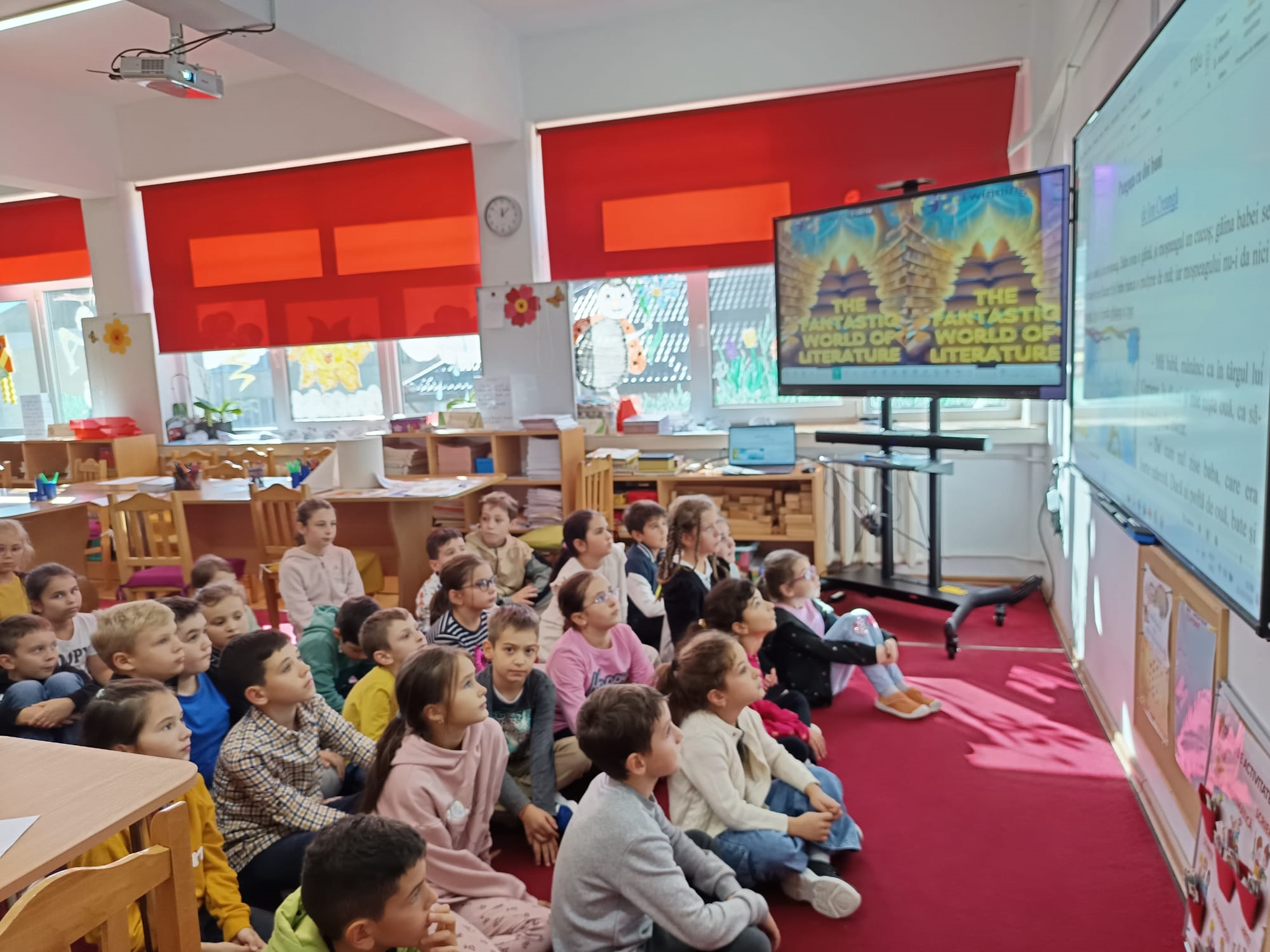 Într-o sală de clasă modernă, luminoasă, cu jaluzele roșii, mobilier din lemn și materiale educaționale pe rafturi, elevi de școală primară stau pe covorul roșu și privesc spre o tablă interactivă digitală pe care este prezentat un text pentru lecția de lectură, pe un alt panou digital fiind vizibil sigla proiectului eTwinning, cu textul, în limba engleză, „The Fantastic World of Literature” (Lumea fantastică a literaturii)