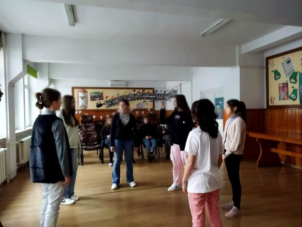 Un grup de elevi se află într-o sală de clasă, stând în picioare, într-o activitate de grup, doi dintre ei find implicați într-o activitate mai apropiată, iar in fundal sunt alți elevi, așezați pe scaune, urmărind scena