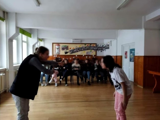 Într-o sală de clasă, un elev este într-o interacțiune mai apropiată cu o persoană adultă în cadrul unui unui exercițiu didactic, iar în fundal sunt alți elevi, așezați pe scaune, urmărind scena