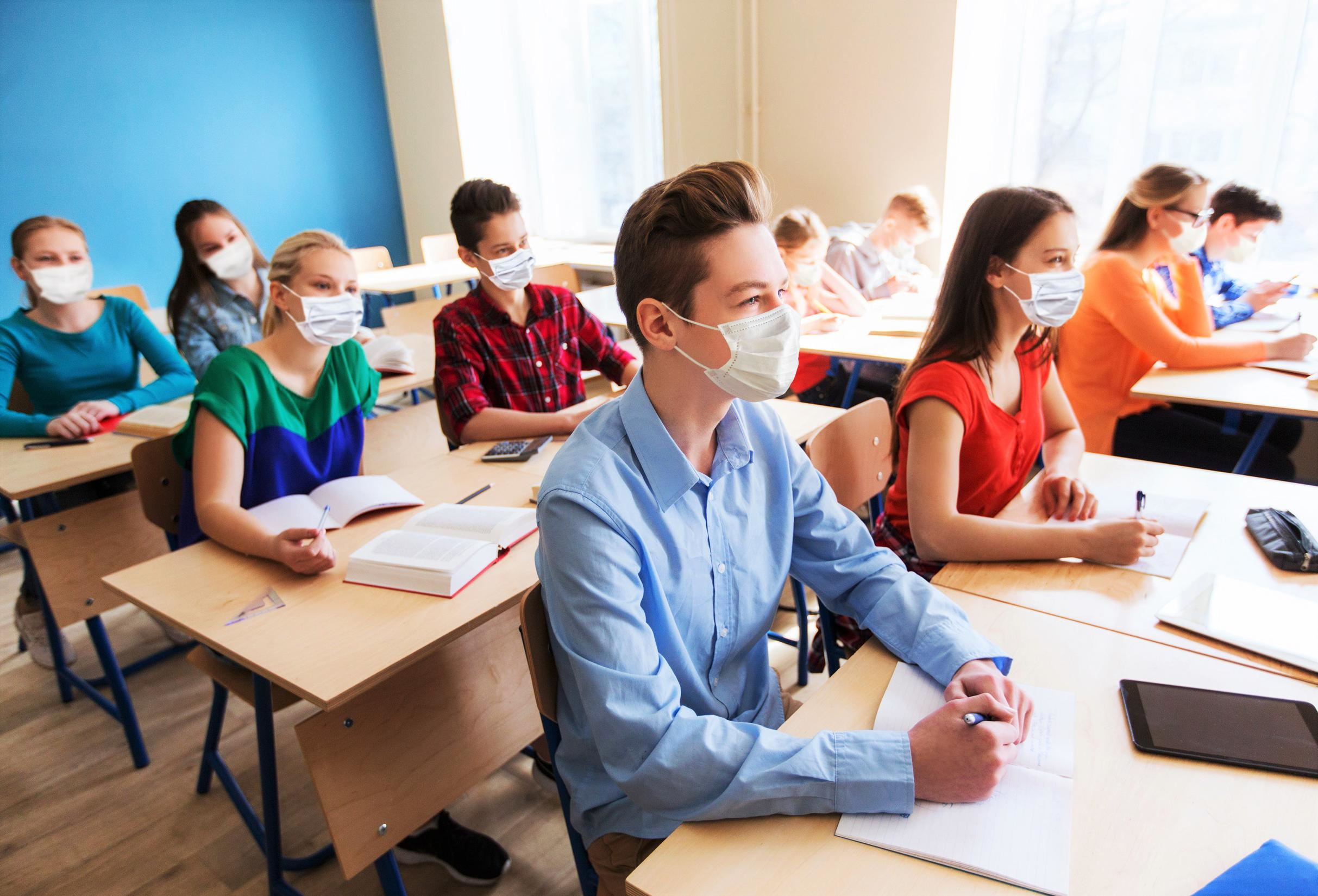 Un grup de elevi, purtând măști medicale de protecție, sunt așezați în bănci într-o sală de clasă luminoasă, fiecare cu câte o carte sau caiet în față, unii scriind, alții fiind concentrați la lecție